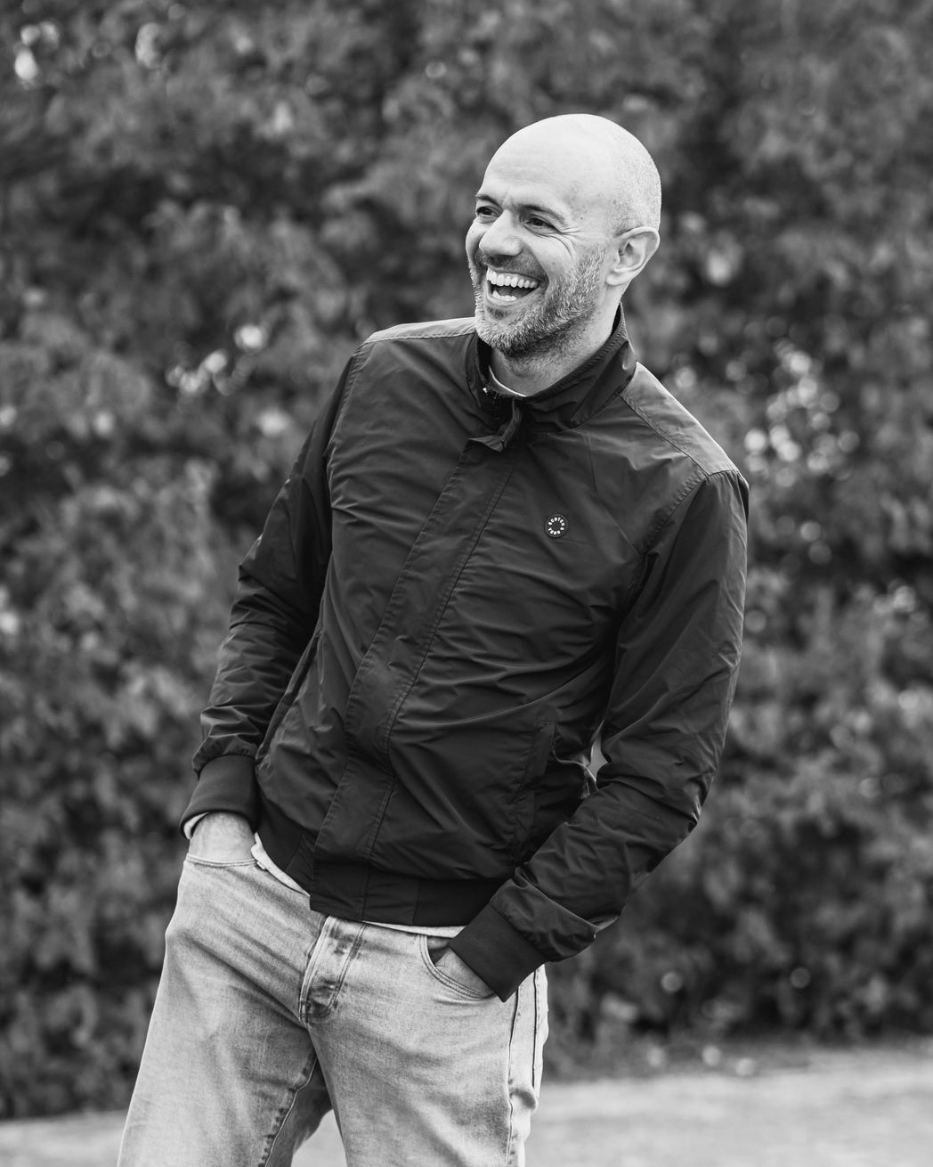 Black and white portrait of Kris Van Herzeele laughing outdoors, hands in the pockets of a dark jacket.