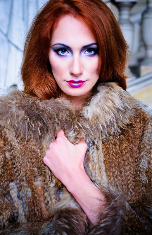 A red-haired woman in a fur coat looks into the camera, violet make-up against cool stone.