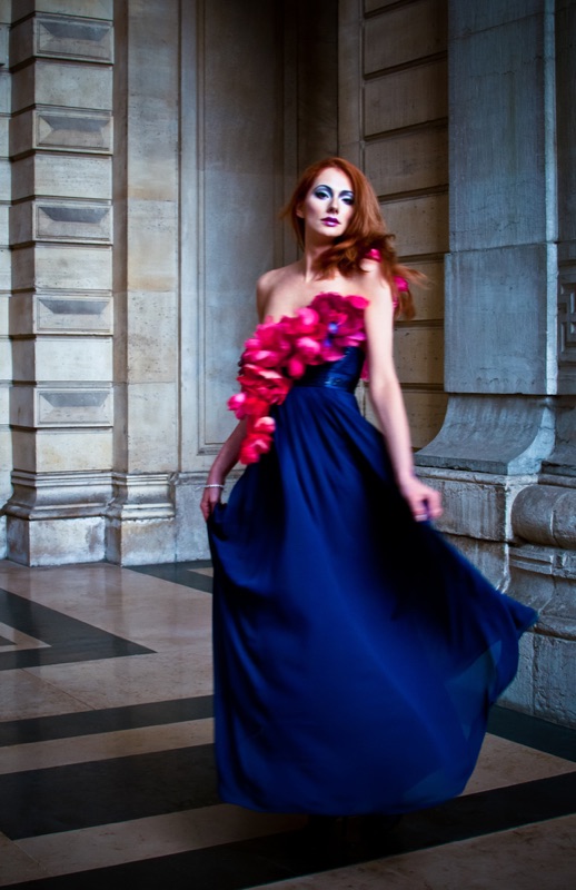 A red-haired woman in a long blue gown with red ruffles moves through a stone colonnade.