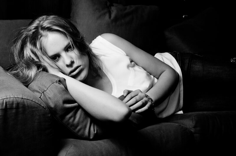 Black and white: a blonde woman rests her head on a dark sofa cushion and looks into the camera.