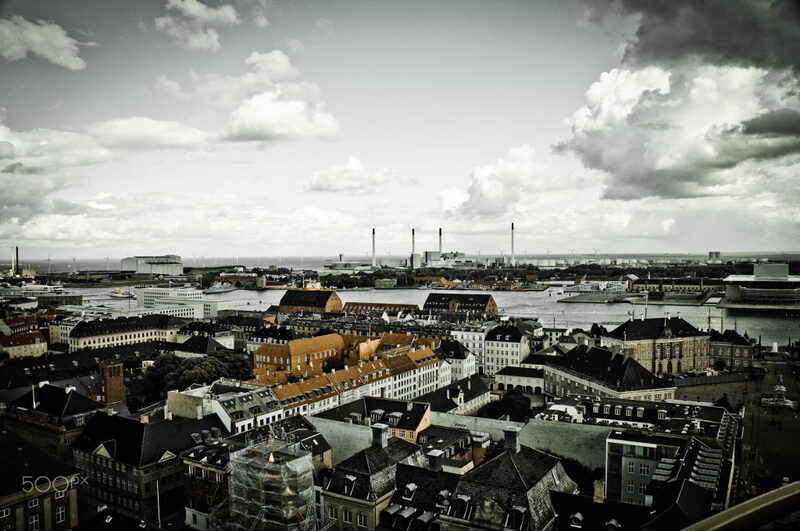 Copenhagen rooftops stretch towards the harbour and chimneys under a bright, cloudy sky.
