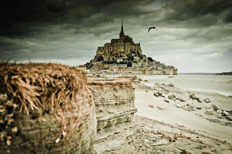 Mont-Saint-Michel under a heavy grey sky, seen across the tidal flats with a bird overhead.