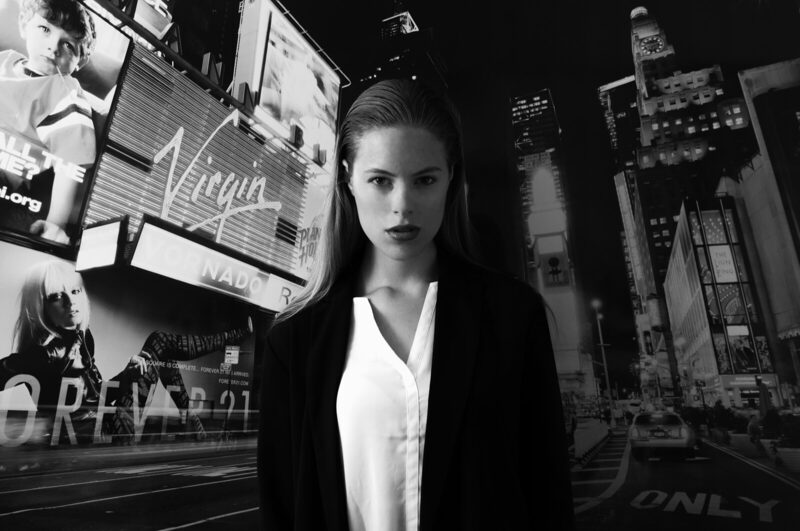 Black and white: a young woman in a dark coat stands in front of a Times Square backdrop.