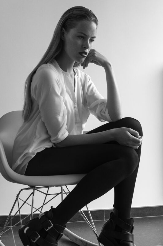 Black and white: a young woman sits on a white chair, chin resting on her hand.